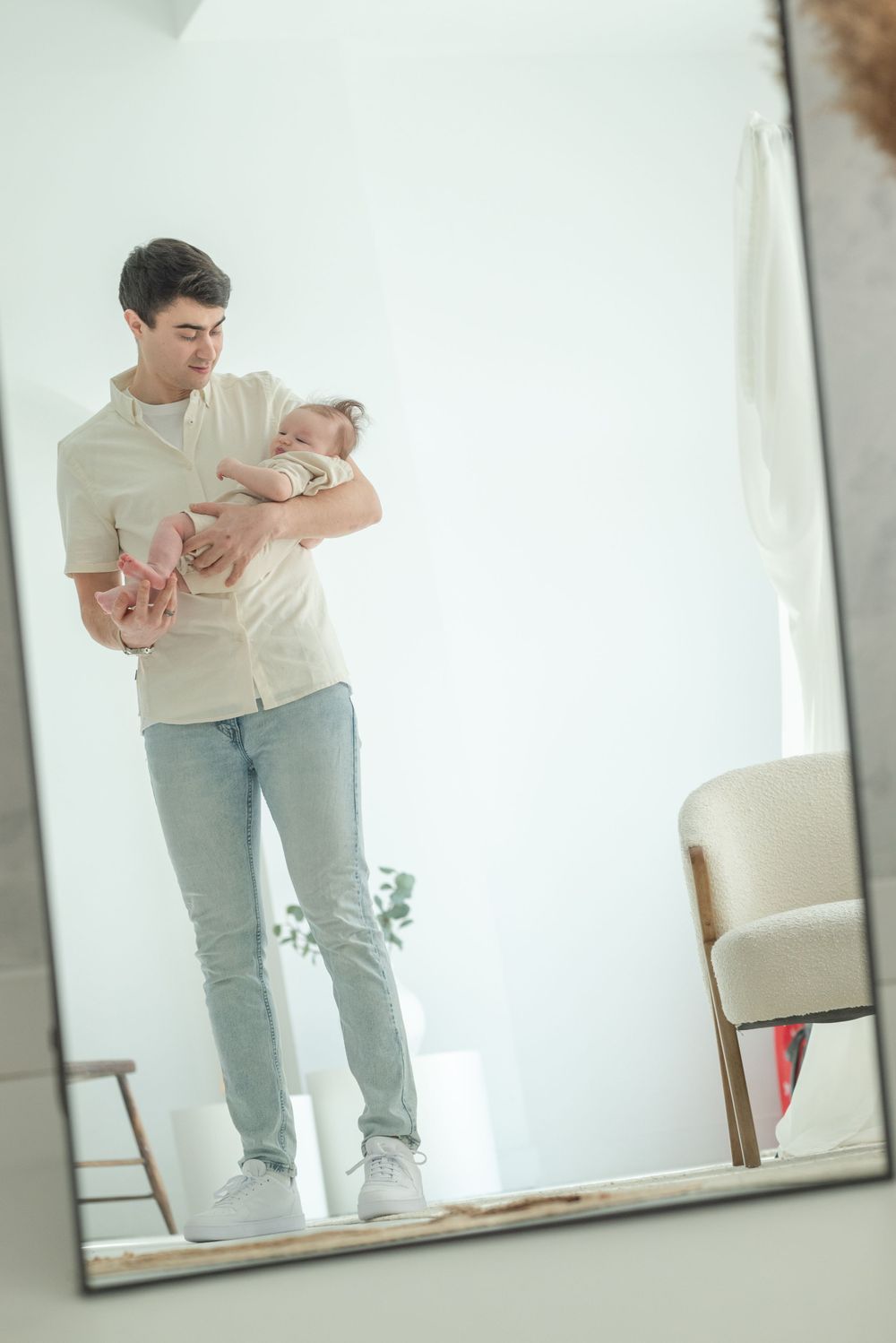 Mirror reflection shows parent holding baby in minimalist white room.