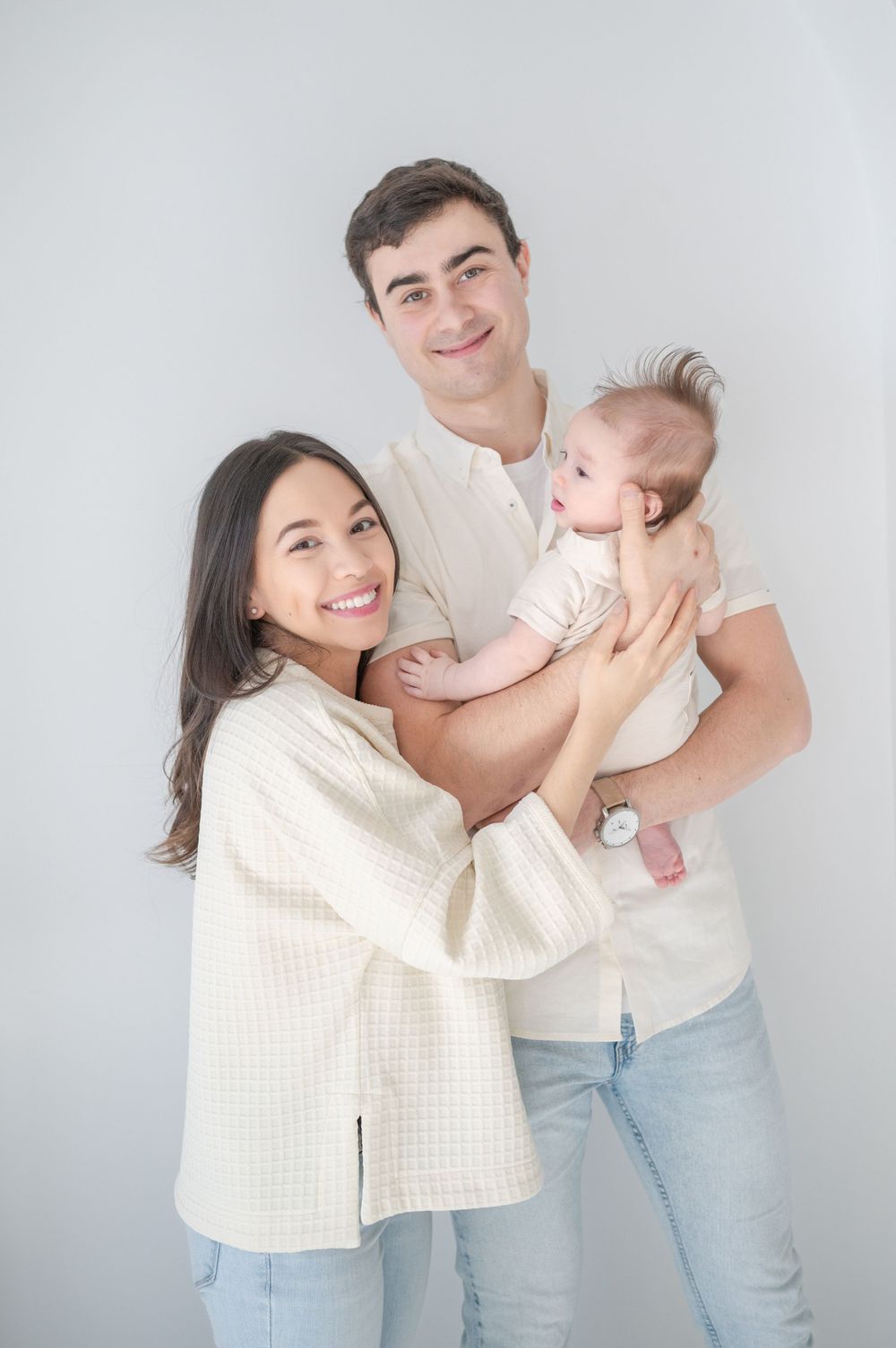 A family of three poses together in matching light-colored outfits against a gray backdrop.