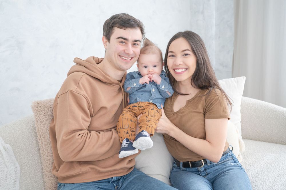 Family portrait on white couch with baby holding teddy bear.