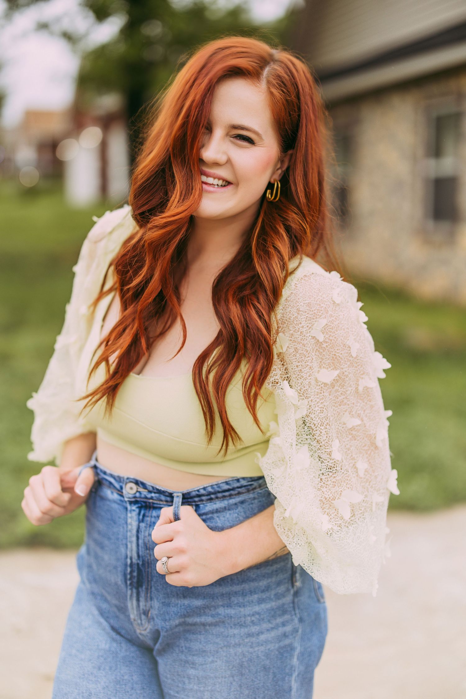 Casual portrait featuring long wavy red hair and lace detailed cream top against outdoor backdrop.