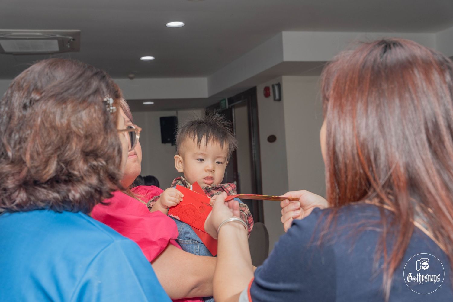 Adults assist in feeding a toddler wearing red during a gathering indoors.