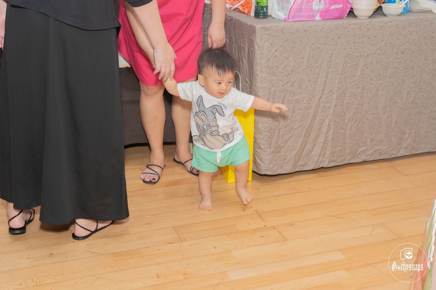 Small child in casual attire walking on hardwood floor beside adult legs.