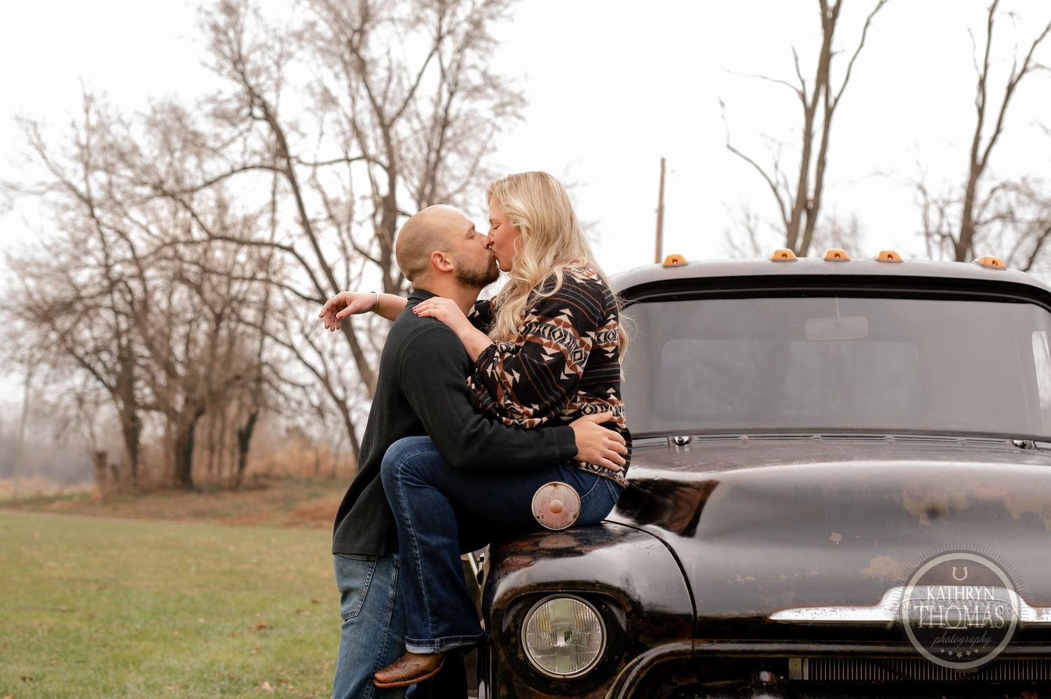 A romantic moment captured between a couple posed on a vintage truck in a rural field.