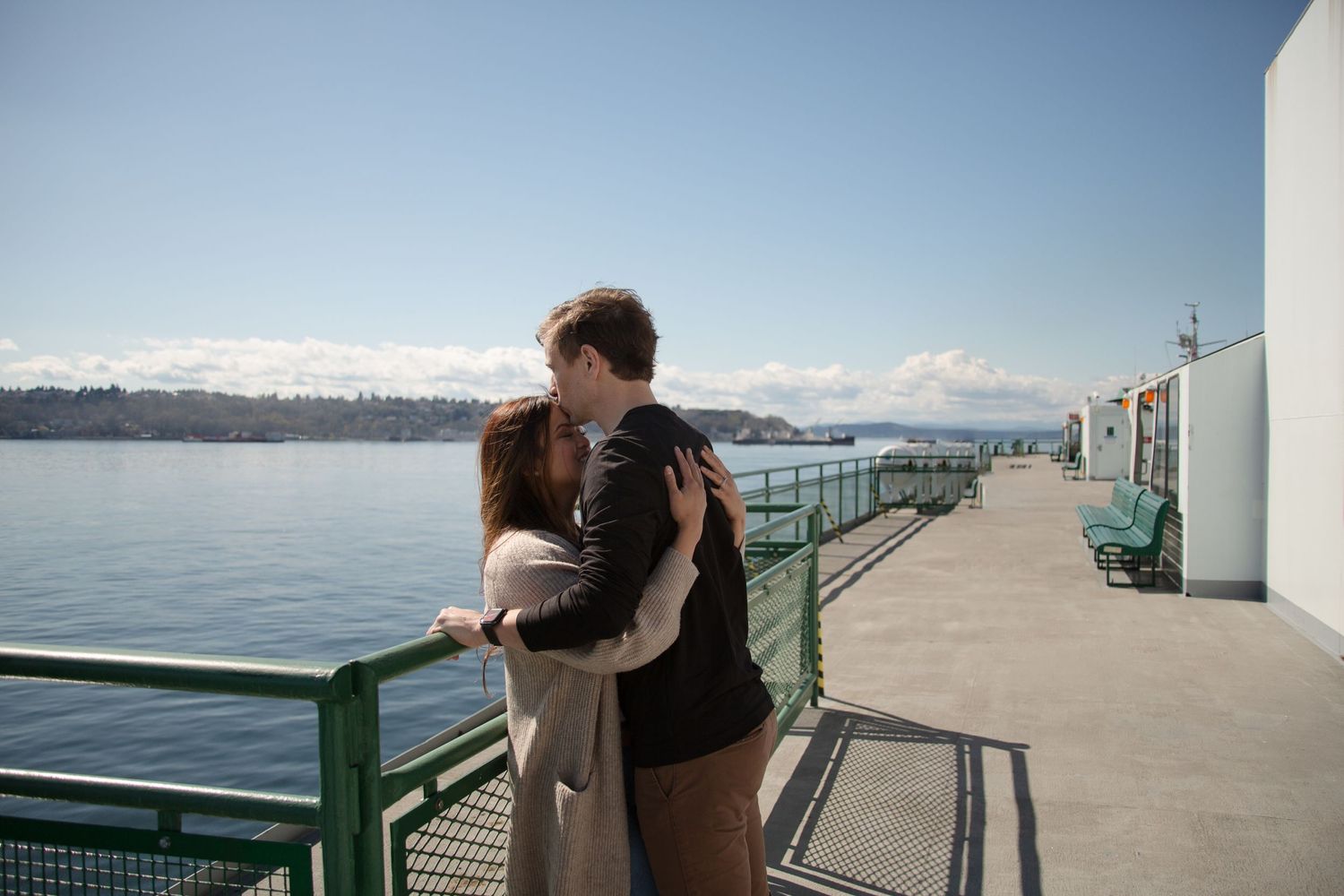 Couple embraces on a waterfront pier with mountains and water in the background on a sunny day.
