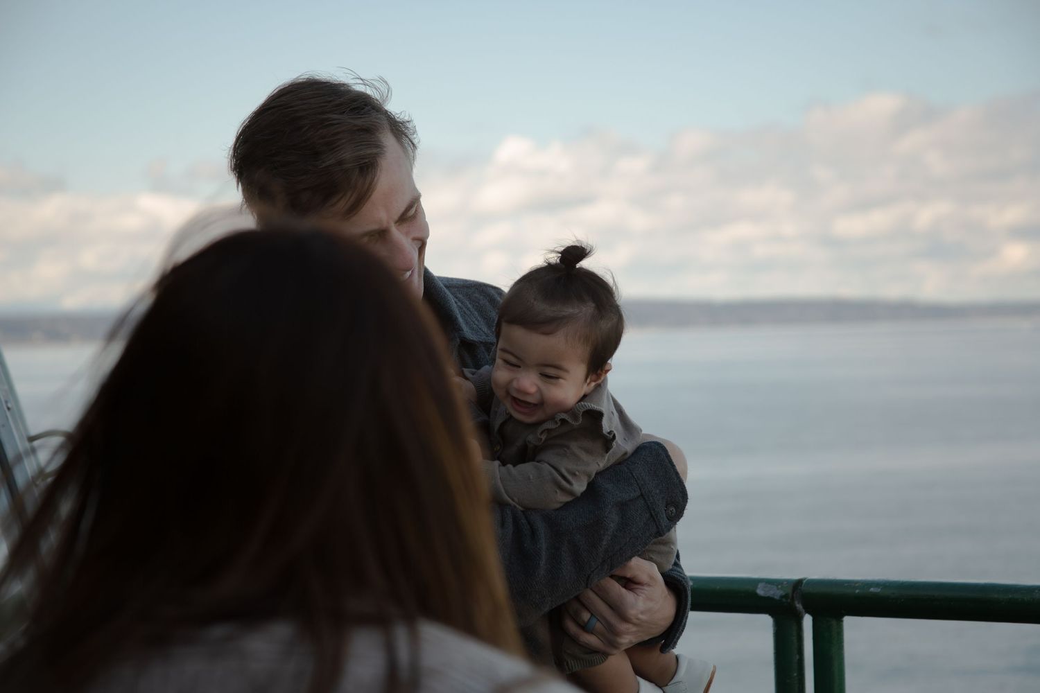 Baby bundled in winter coat held by adults on waterfront overlook at dusk.