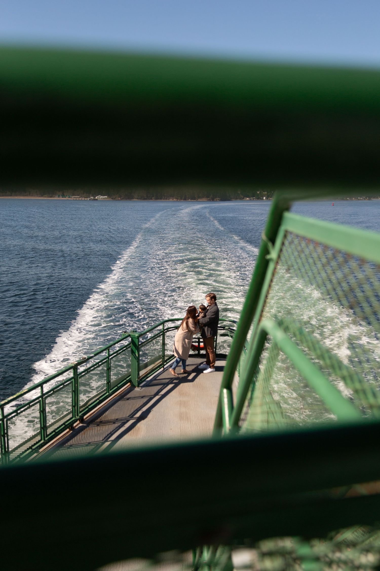 View through railing of couple standing on ferry deck with wake trailing behind vessel.