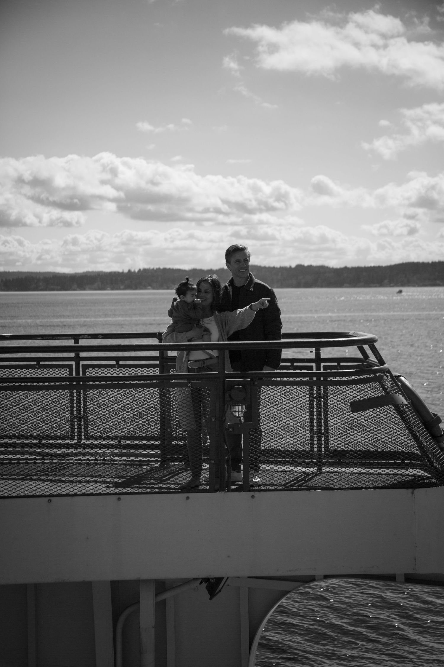 A couple stands on ferry deck overlooking water and distant shoreline in black and white.