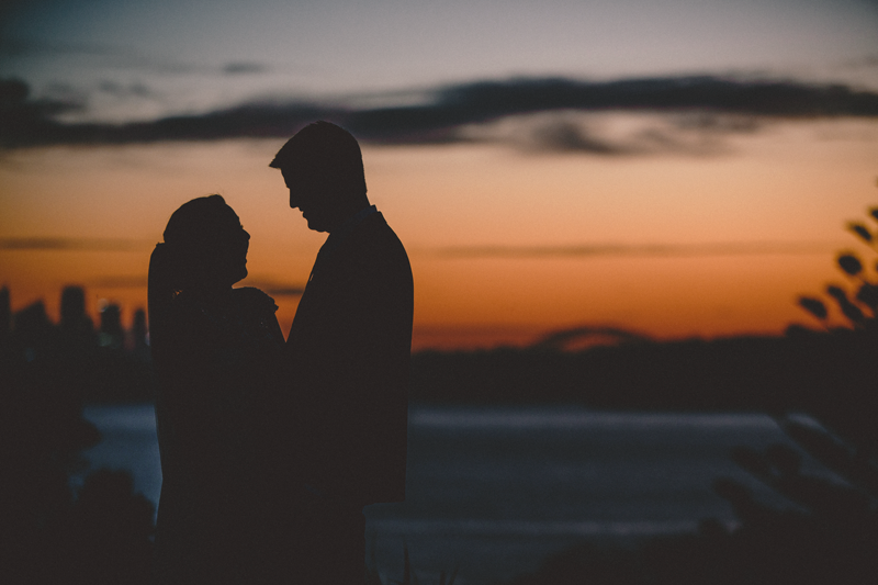Silhouette of couple standing close together against vibrant orange sunset sky with city skyline in background.