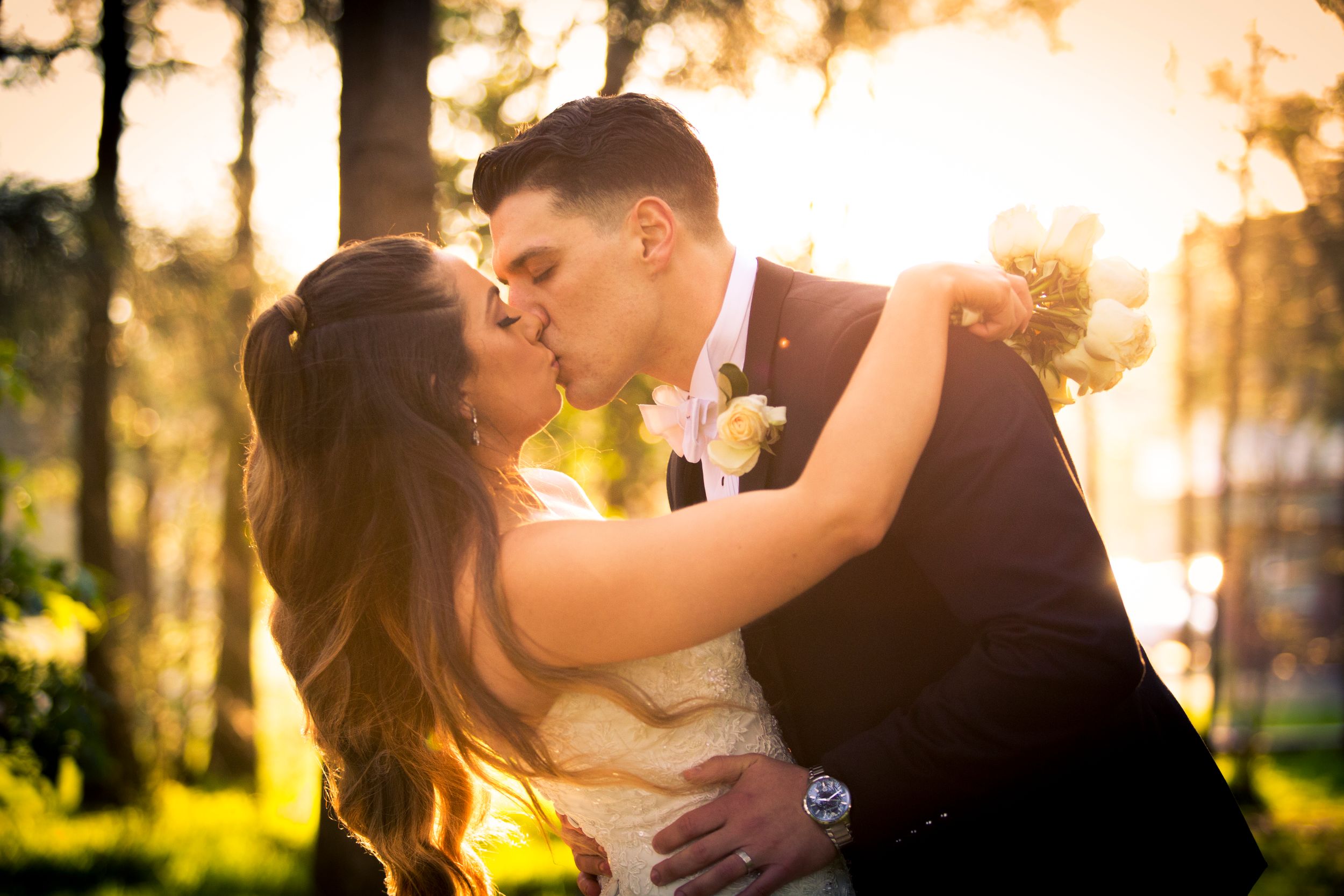 Romantic wedding couple sharing a kiss in golden sunlight filtering through trees at sunset.