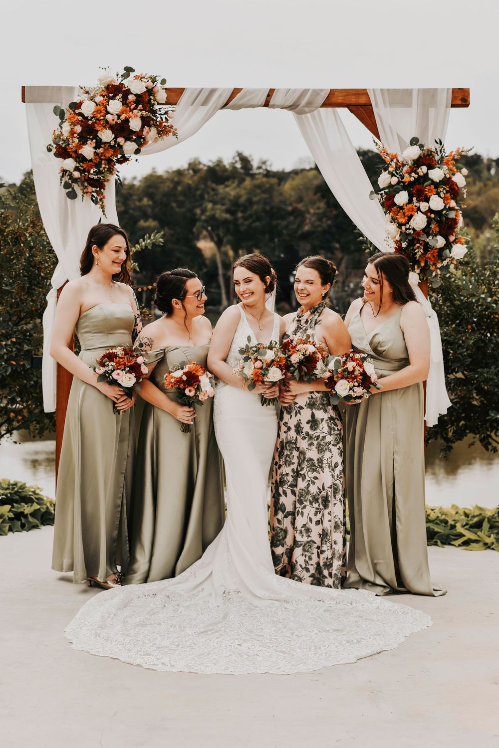 a bride laughs as her bridal party smile at her as they stand in front of their altar which is draped in white drapery