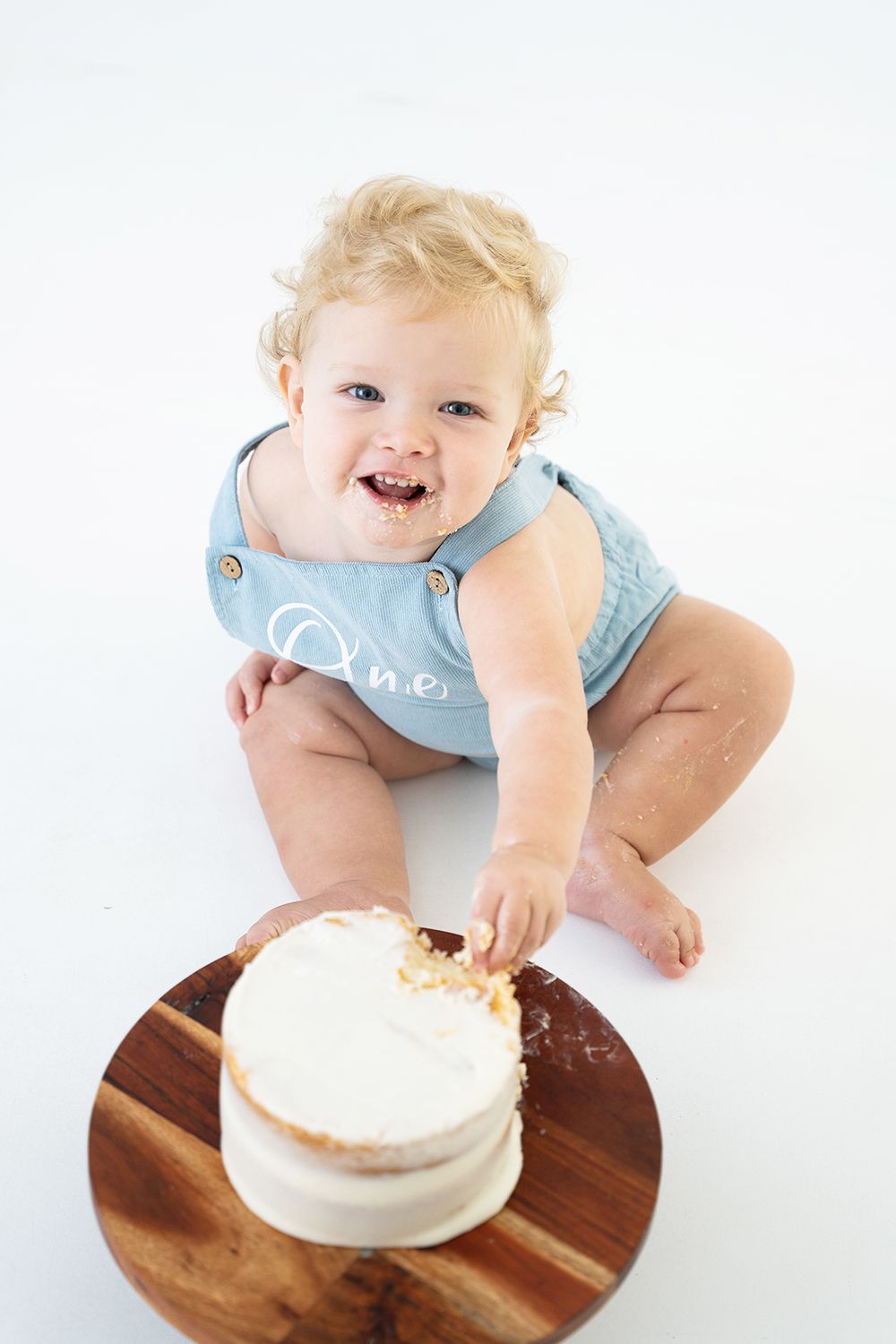 Happy baby in light blue outfit reaches for white cake on wooden serving board against white background.
