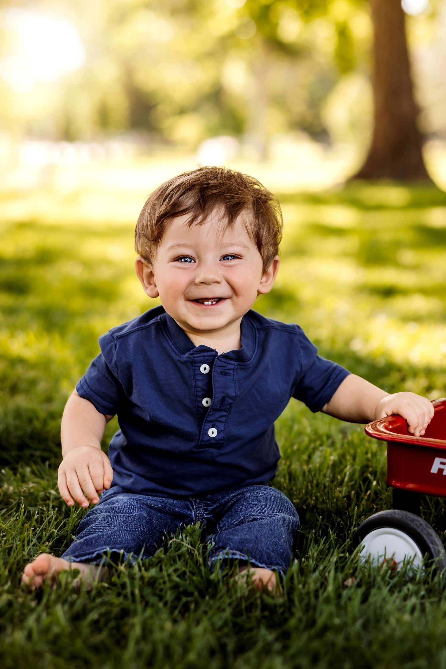 A happy child in a navy blue outfit sits on green grass during a sunny outdoor photo session.
