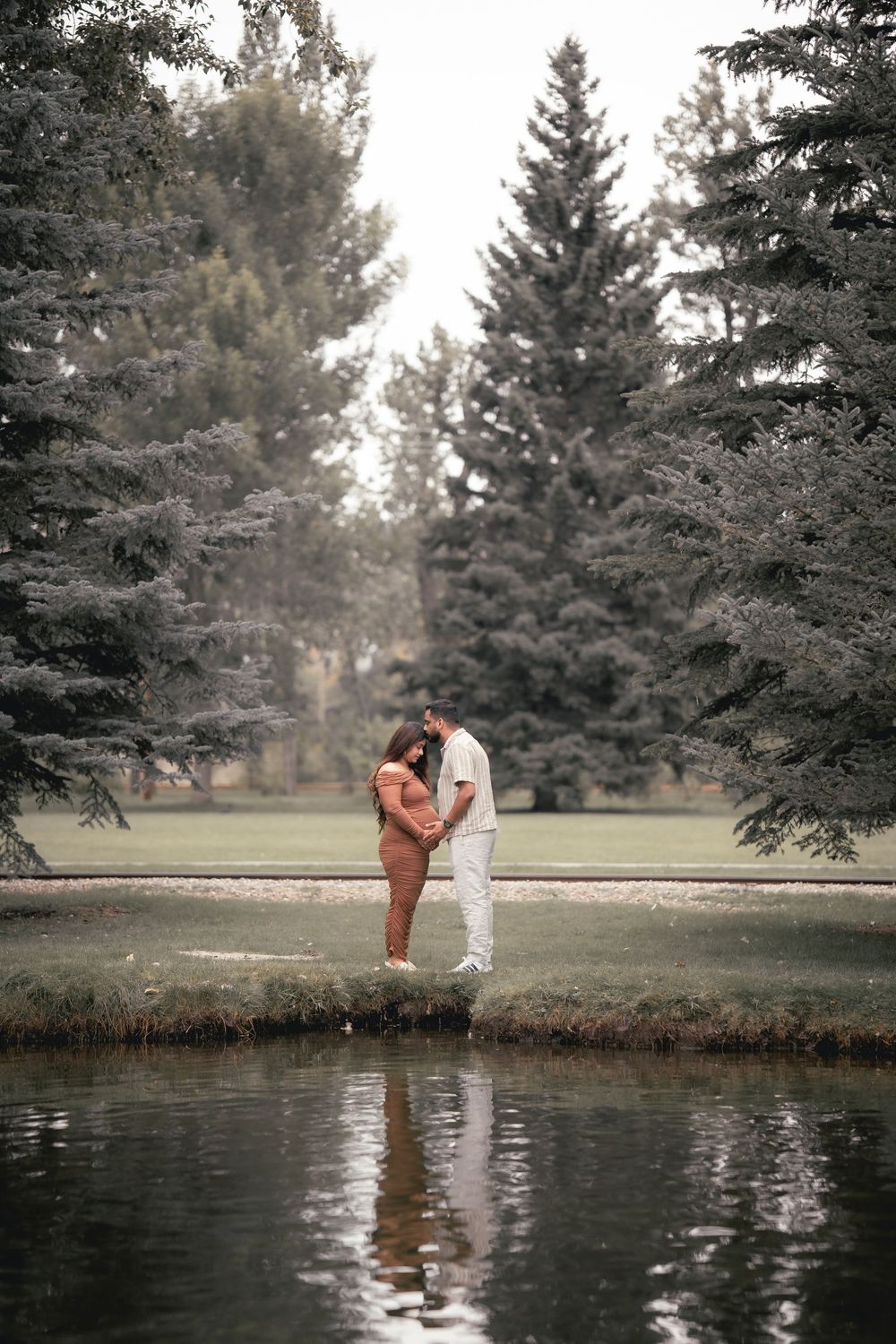 A romantic couple stands by a tranquil pond surrounded by evergreen trees in a serene outdoor setting.