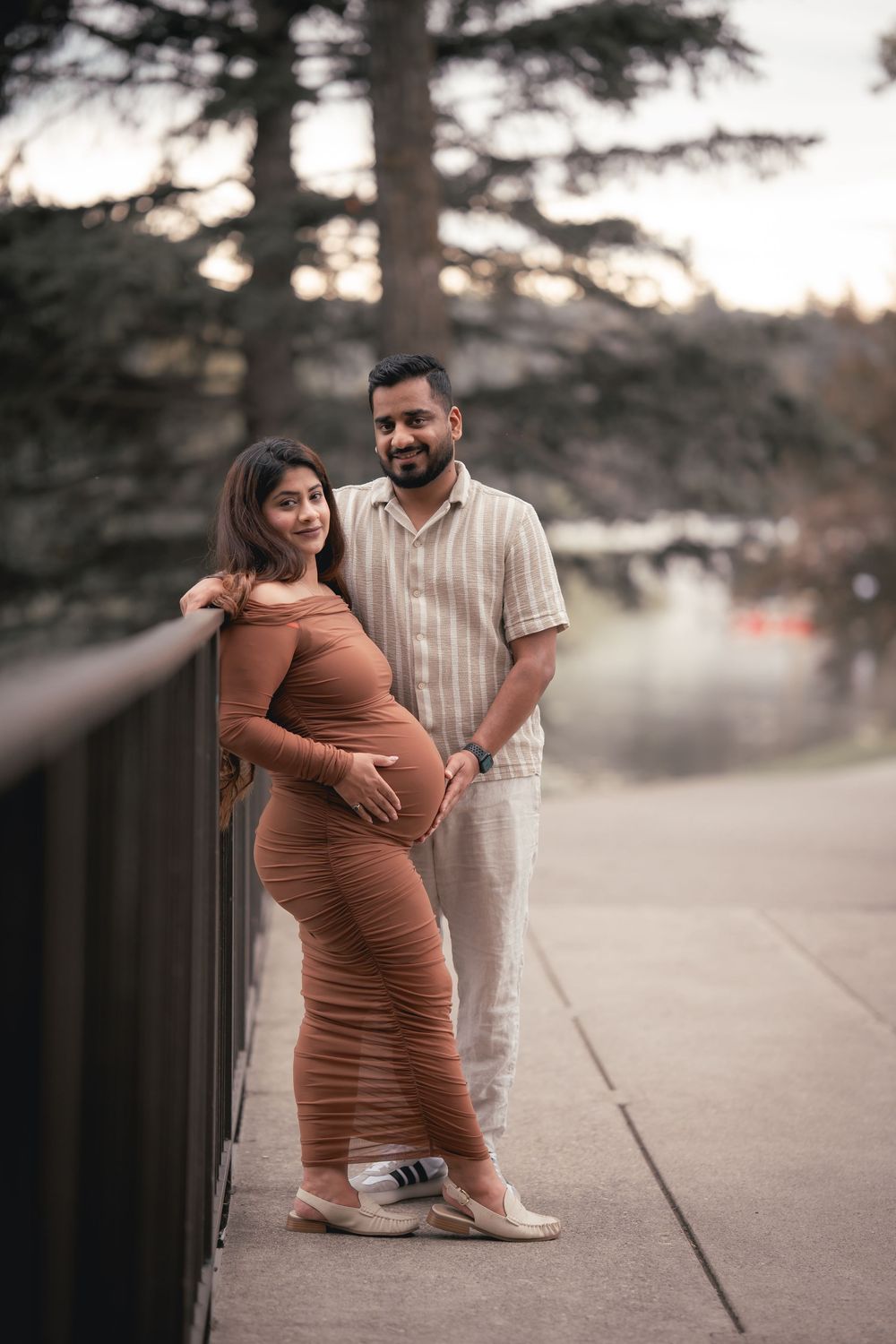 A couple poses outdoors during a maternity photoshoot at sunset with the expectant mother wearing a brown dress.