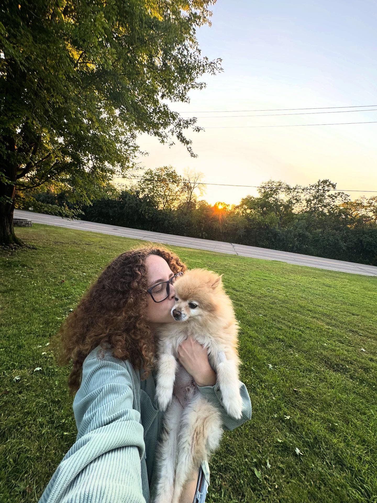A person in a mint sweater embraces a fluffy white dog outdoors during sunset in a grassy field.