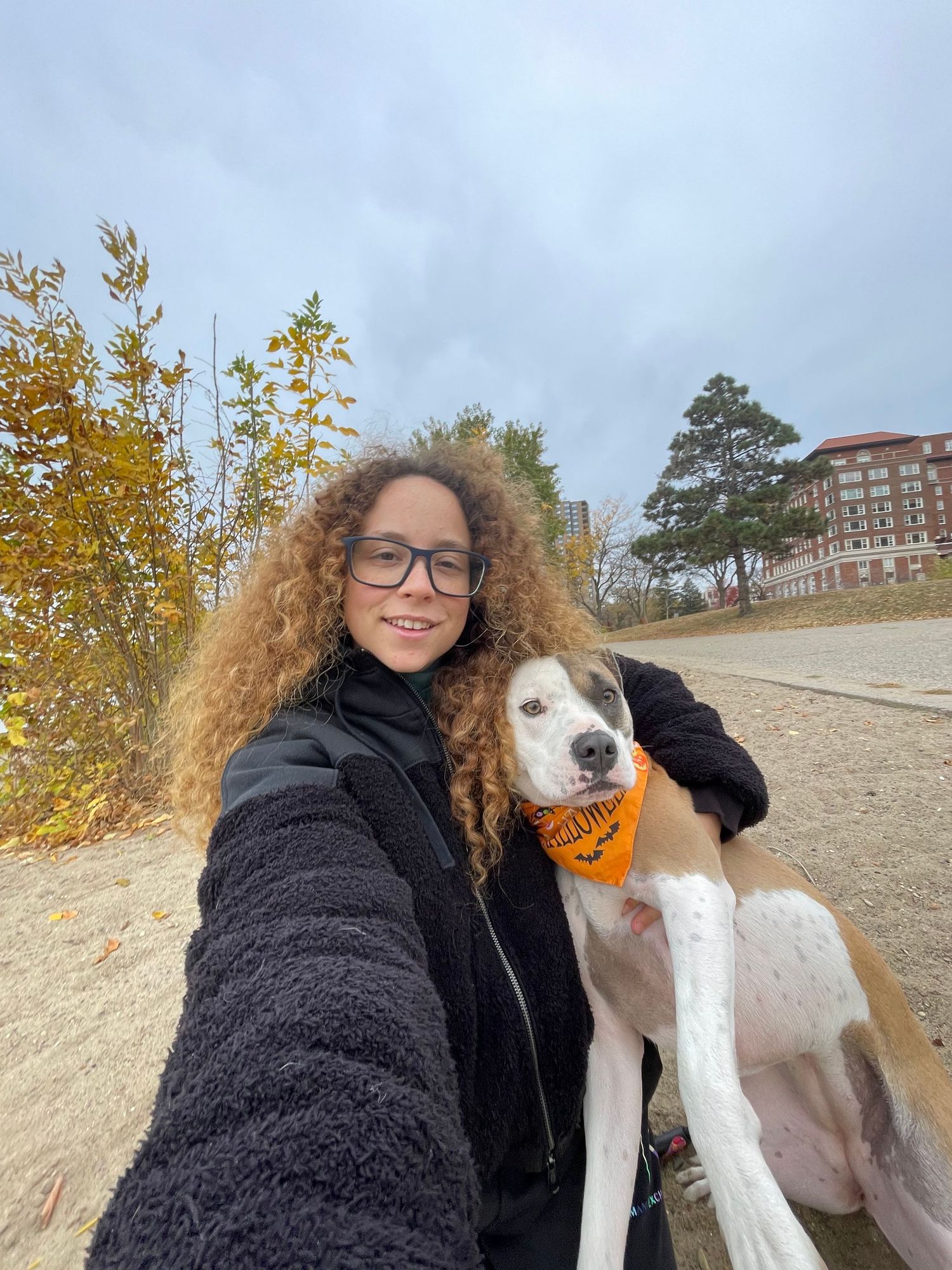 A person in a black winter coat takes selfies with a friendly white and spotted dog against an autumn outdoor backdrop.
