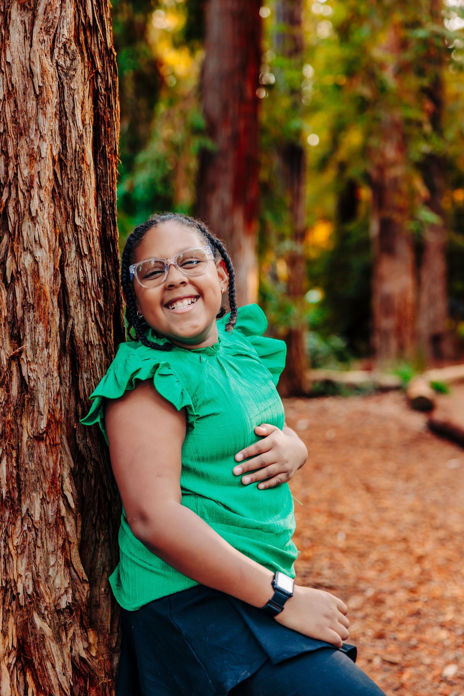 A person wearing a bright green shirt sits among tall redwood trees on a forest floor covered in fallen leaves.