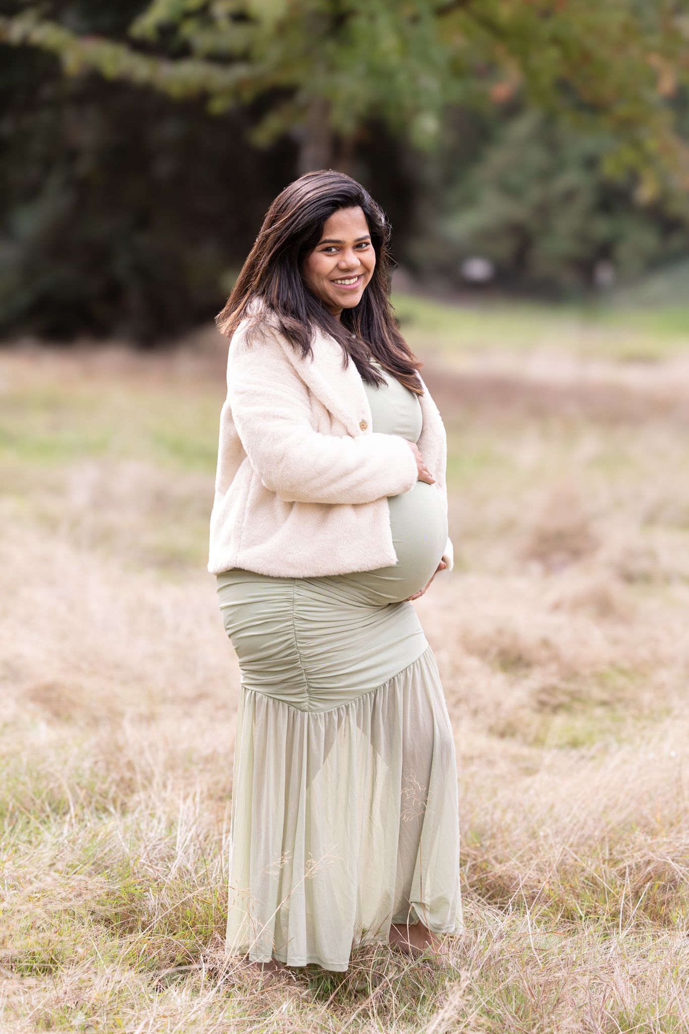 A person in a cream sweater and sage green skirt sits gracefully in a sunlit meadow with tall grass.