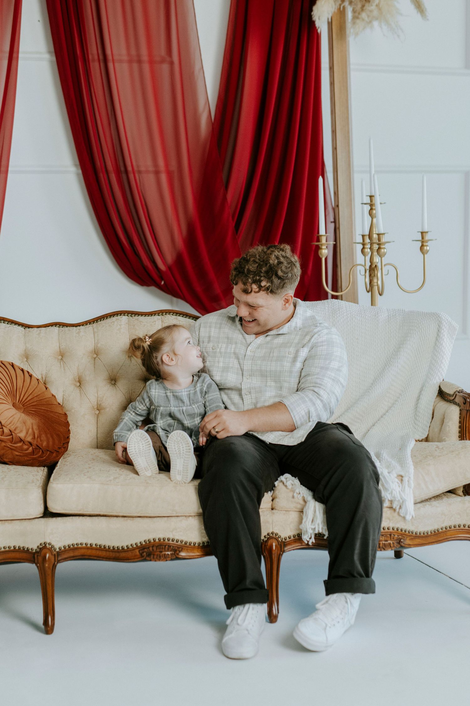 Two people sit together on an elegant cream-colored sofa with red curtains and gold accents in the background.
