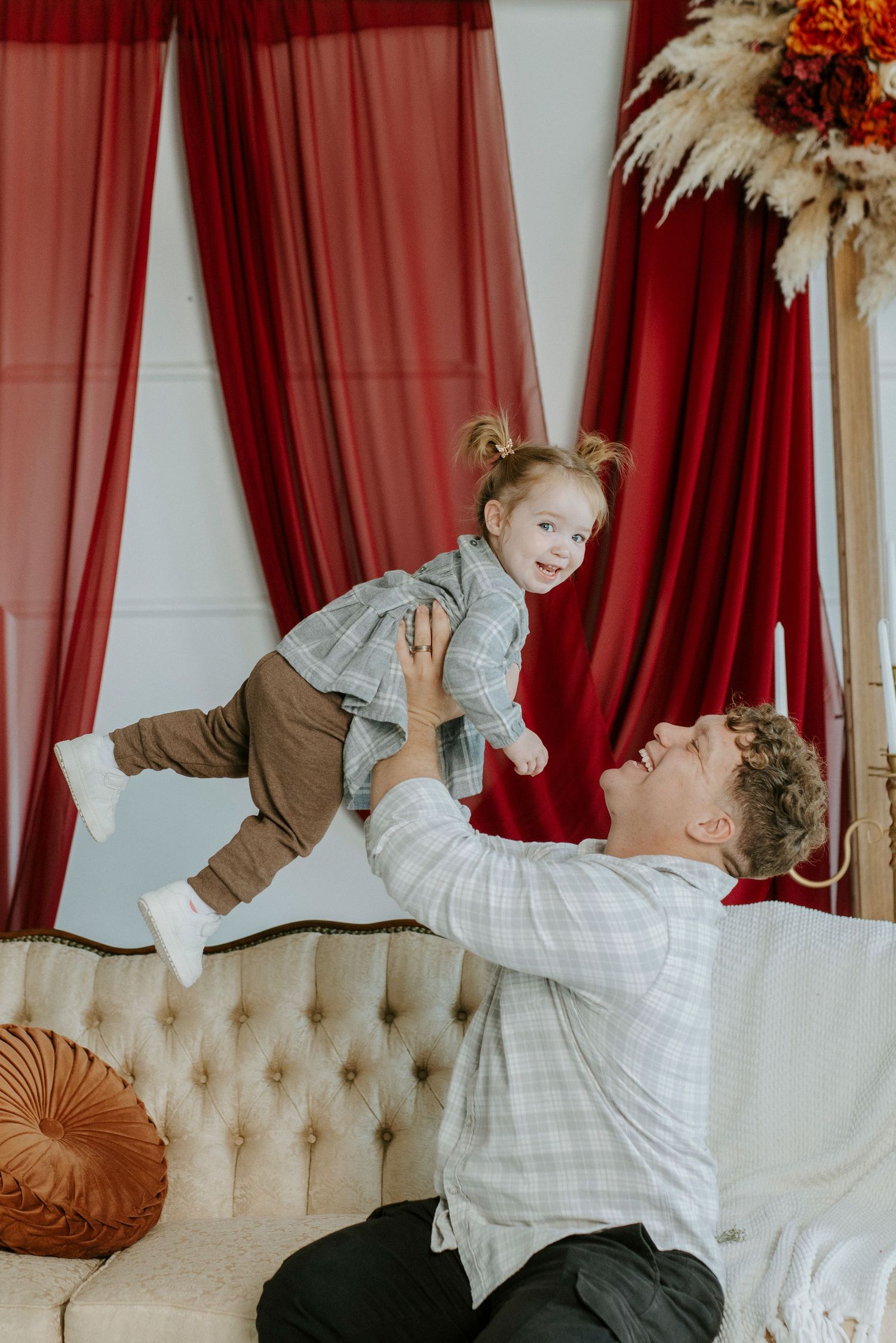 A couple in casual attire shares a playful moment on a beige tufted sofa against red curtains.