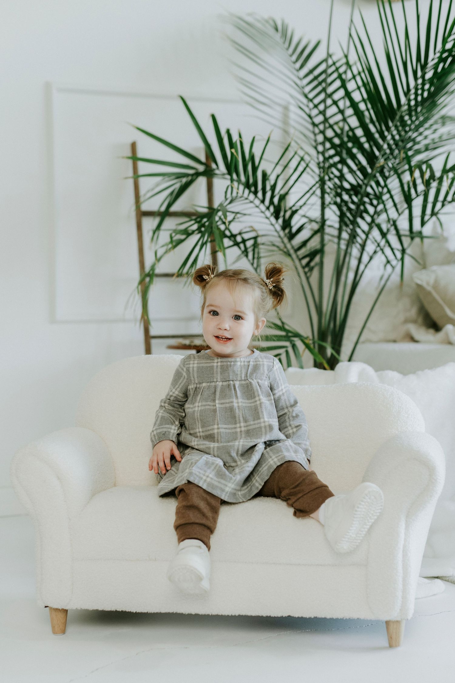A child in a gray plaid shirt and brown pants sits on a white couch with a potted palm plant in the background.