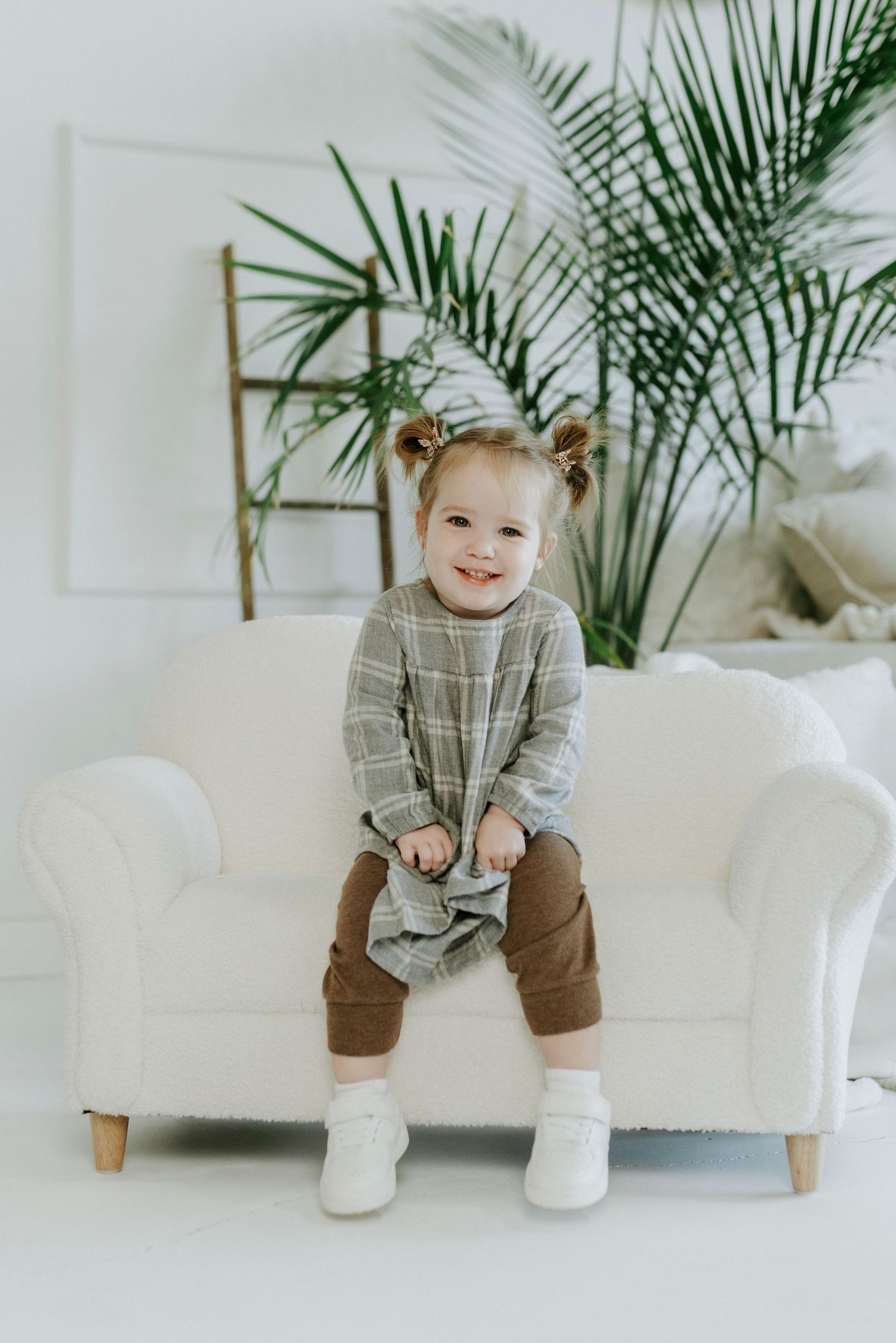 Small child sits on white couch wearing gray sweater and brown pants with indoor palm plant in background.