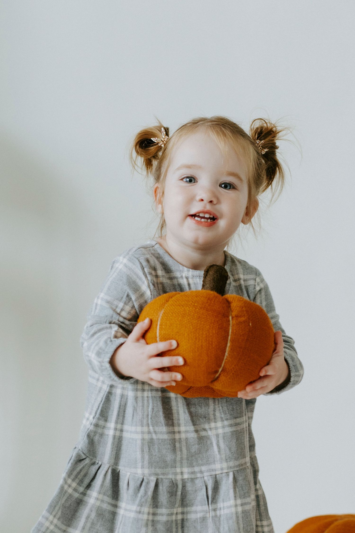 A small child in a gray dress holds a bright orange pumpkin against a white background.