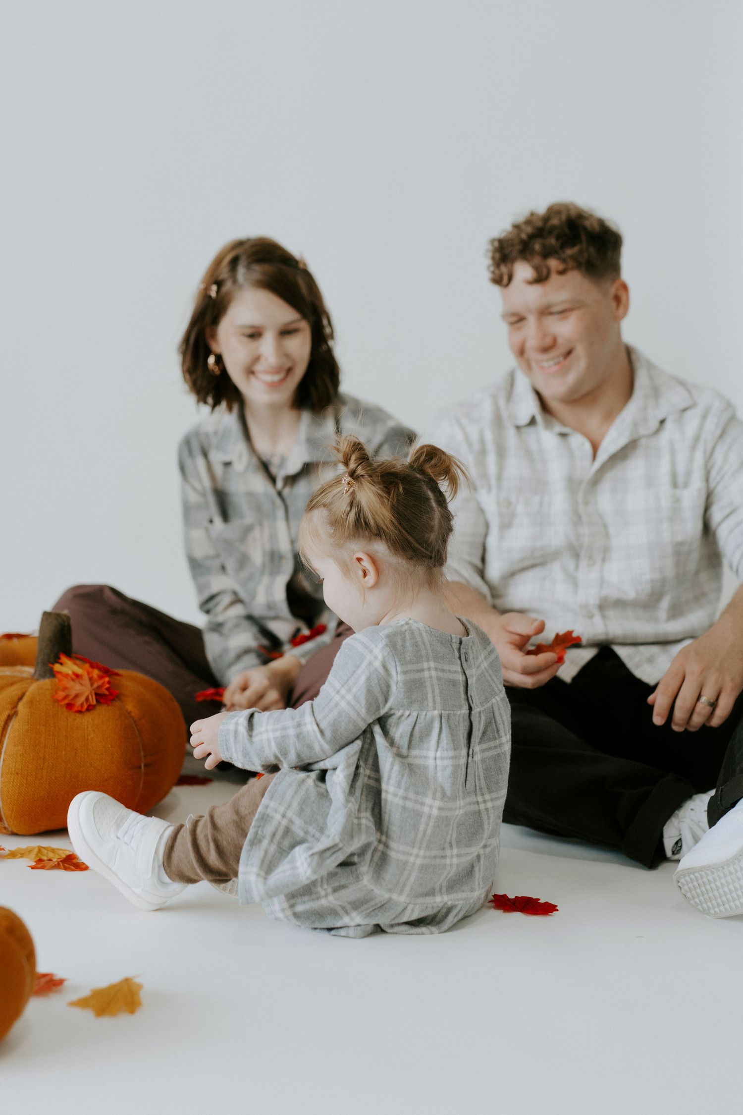 A family sits together on a white floor with orange pumpkins and autumn leaves for a seasonal photoshoot.