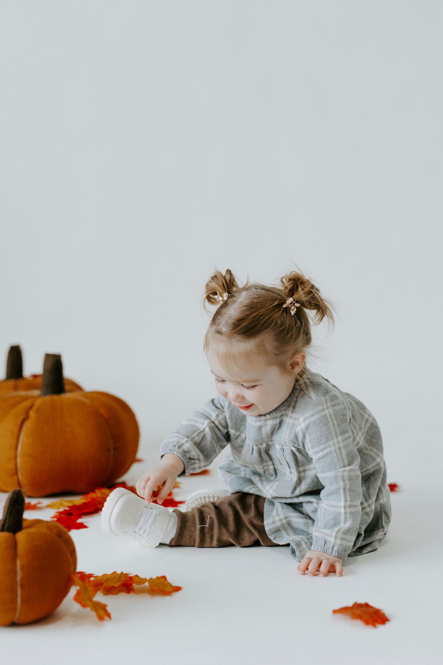 A toddler in gray clothing crawling near orange pumpkins and fall leaves on a white background.
