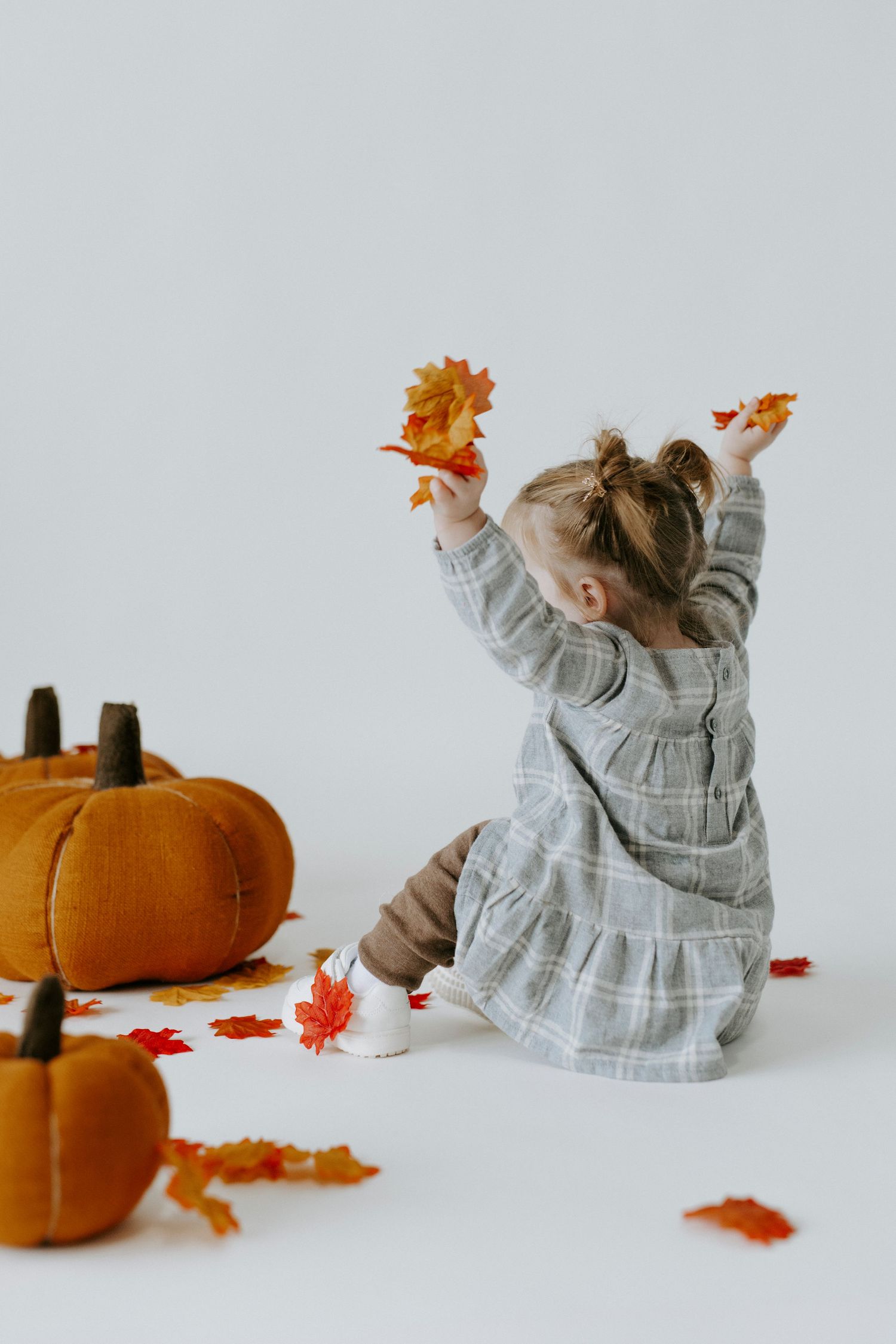Small child in gray dress sits on floor playing with orange autumn leaves and decorative pumpkins against white background.