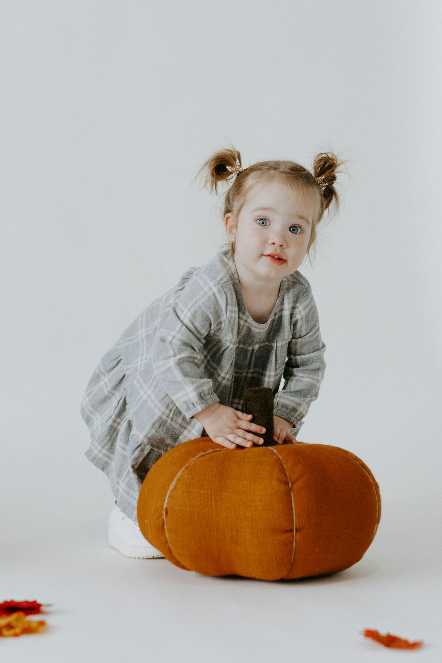 A small child in a gray dress leans over a large orange pumpkin cushion in a minimalist white setting.