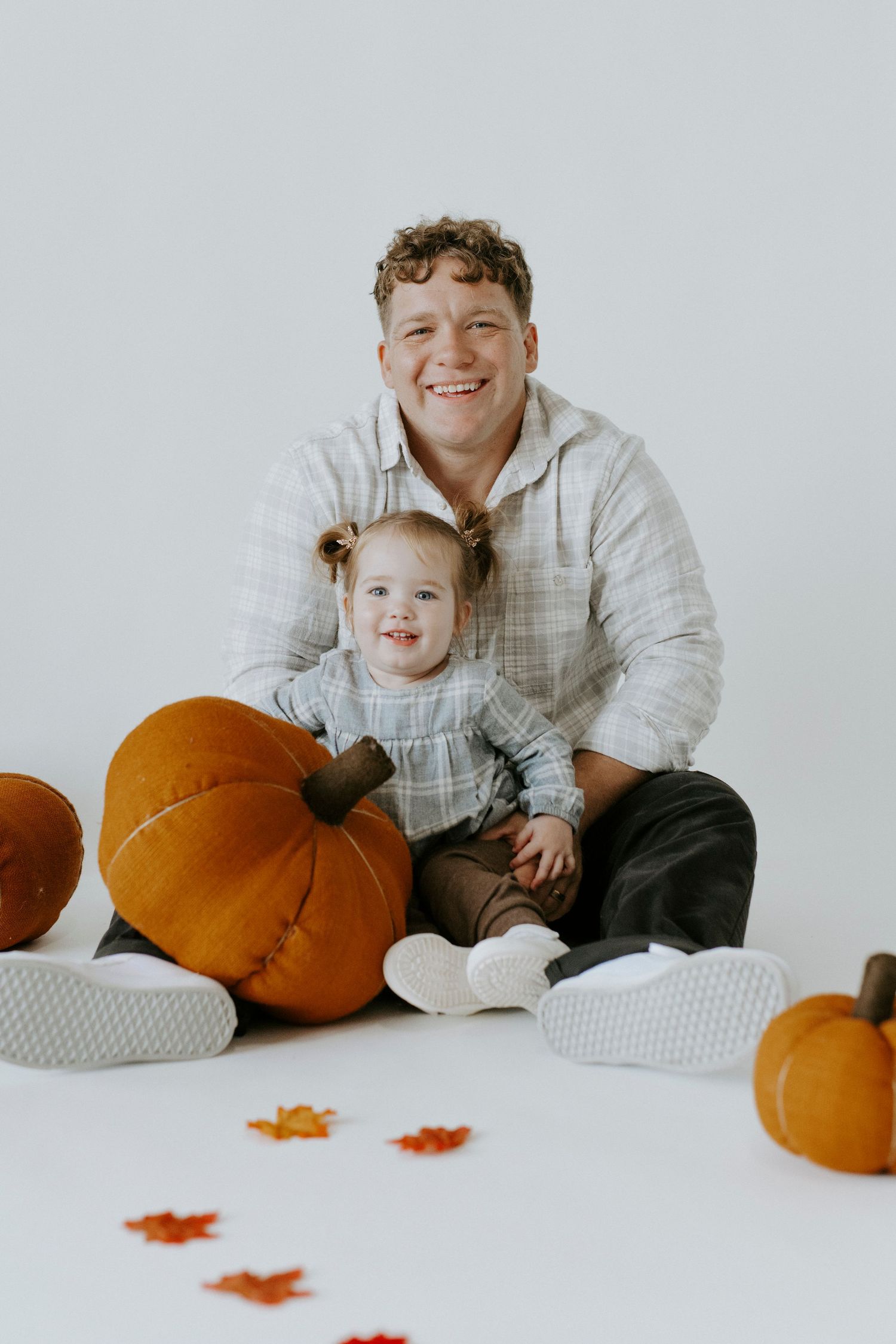 A toddler in a plaid dress sits with pumpkins and fall leaves during an autumn-themed studio photoshoot.