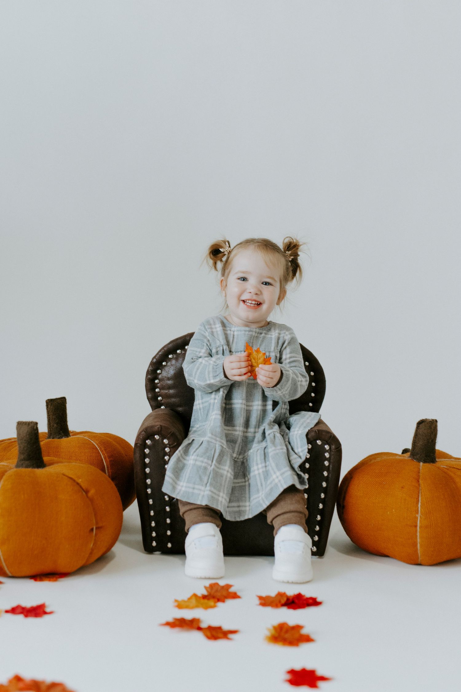A small child sits in a gray chair surrounded by orange pumpkins and red autumn leaves for a fall photo session.