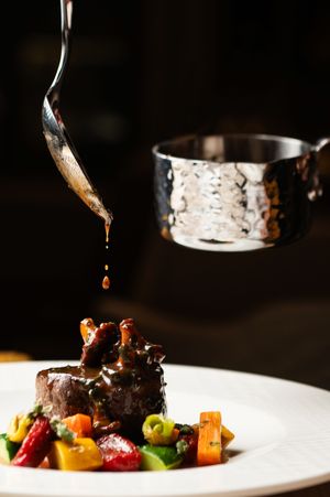 Sauce being drizzled over plated meat and vegetables on white dish.