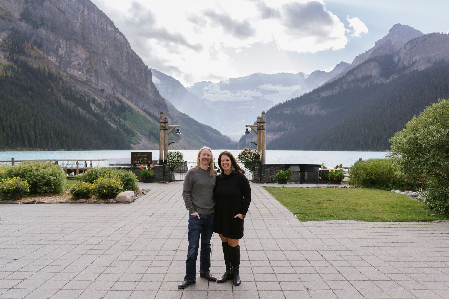 A couple embraces while standing on the rocky shores of Lake Louise in Banff National Park with mountains in the background.