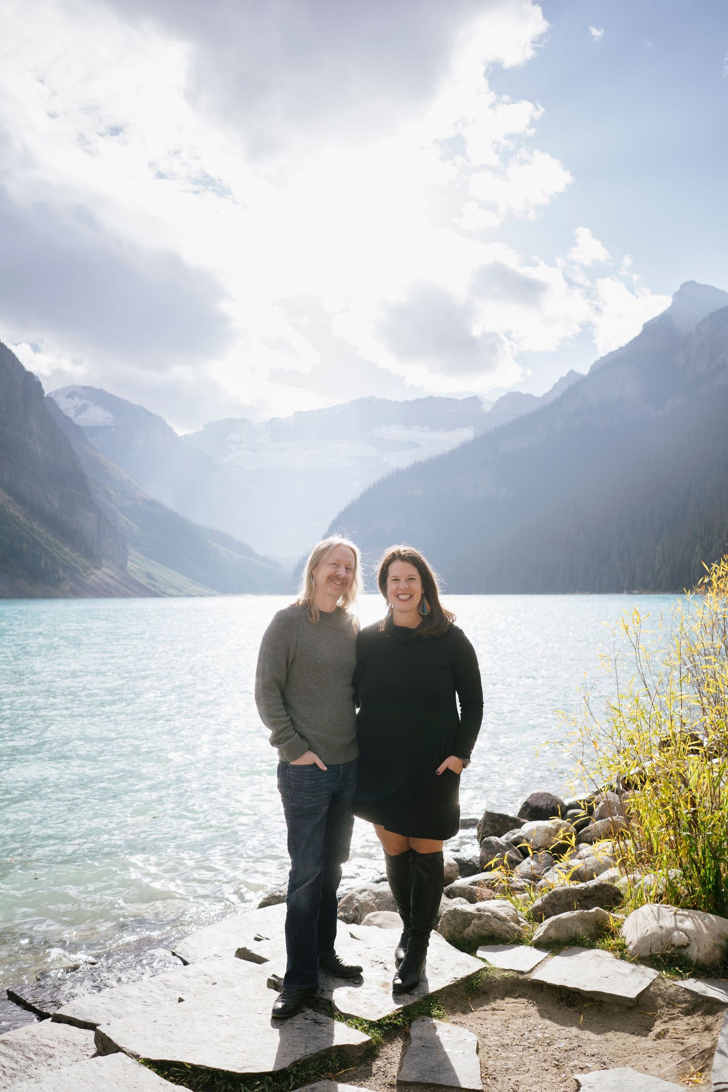 A couple embraces while standing on the rocky shores of Lake Louise in Banff National Park with mountains in the background.