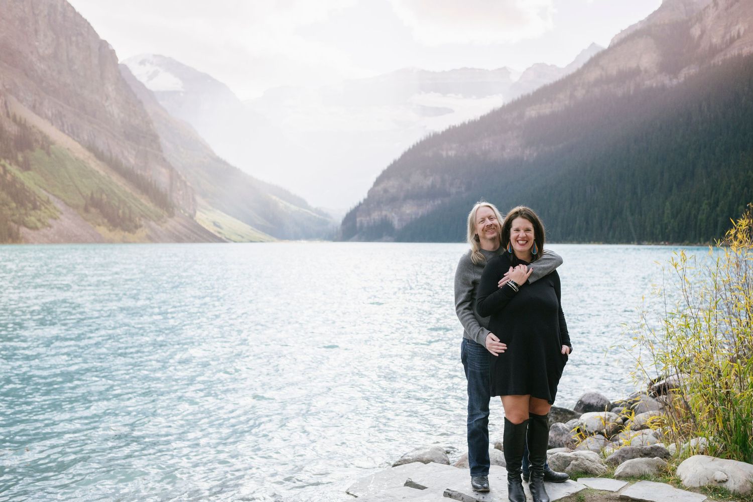 A couple embraces while standing on the rocky shores of Lake Louise in Banff National Park with mountains in the background.