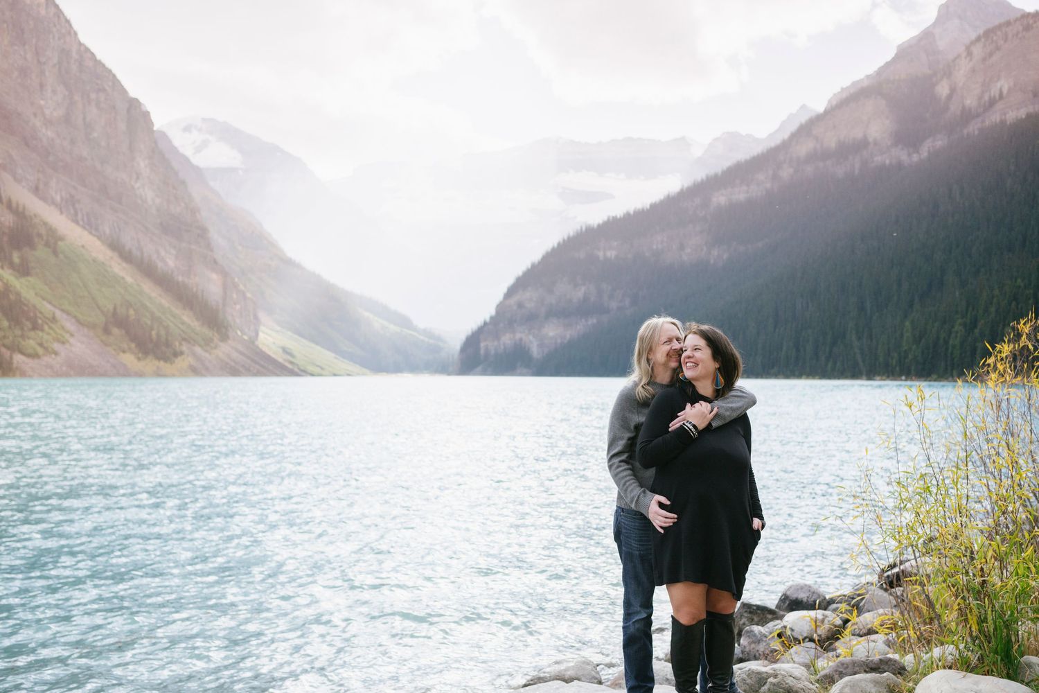 A couple embraces while standing on the rocky shores of Lake Louise in Banff National Park with mountains in the background.