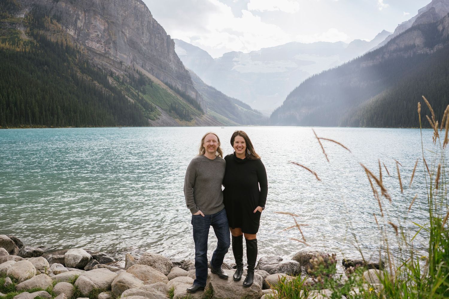 A couple embraces while standing on the rocky shores of Lake Louise in Banff National Park with mountains in the background.