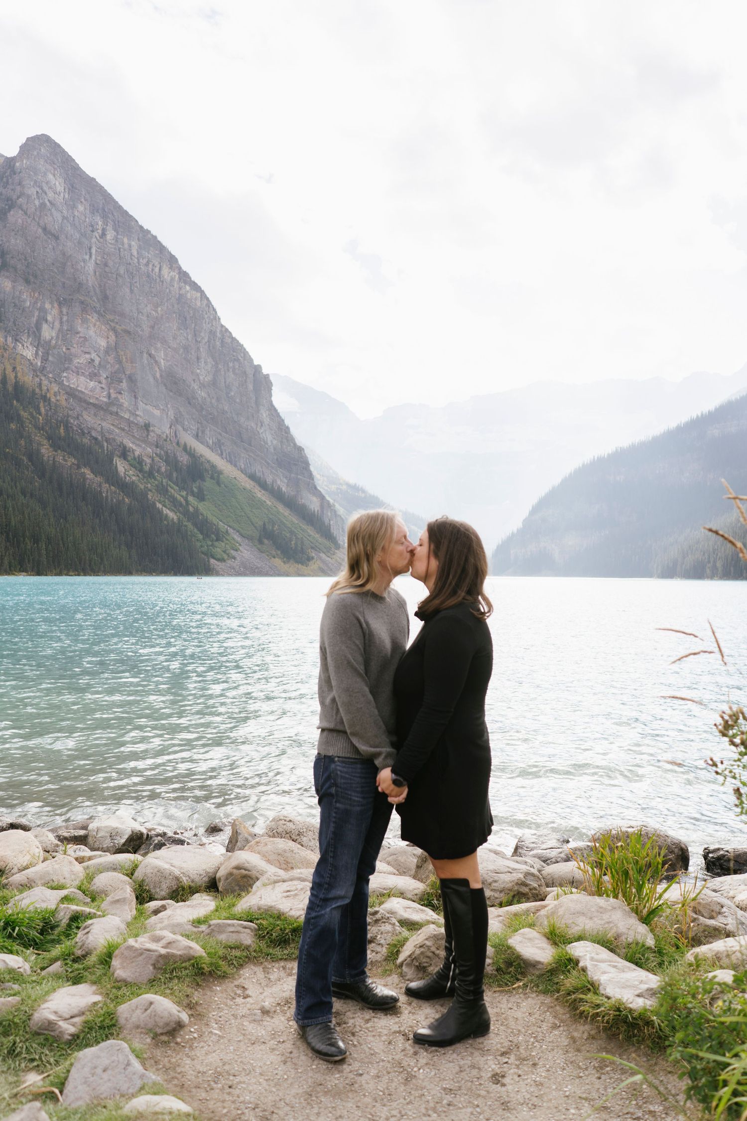 Two people stand together on rocky shoreline overlooking turquoise Lake Louise with mountain peaks in background.