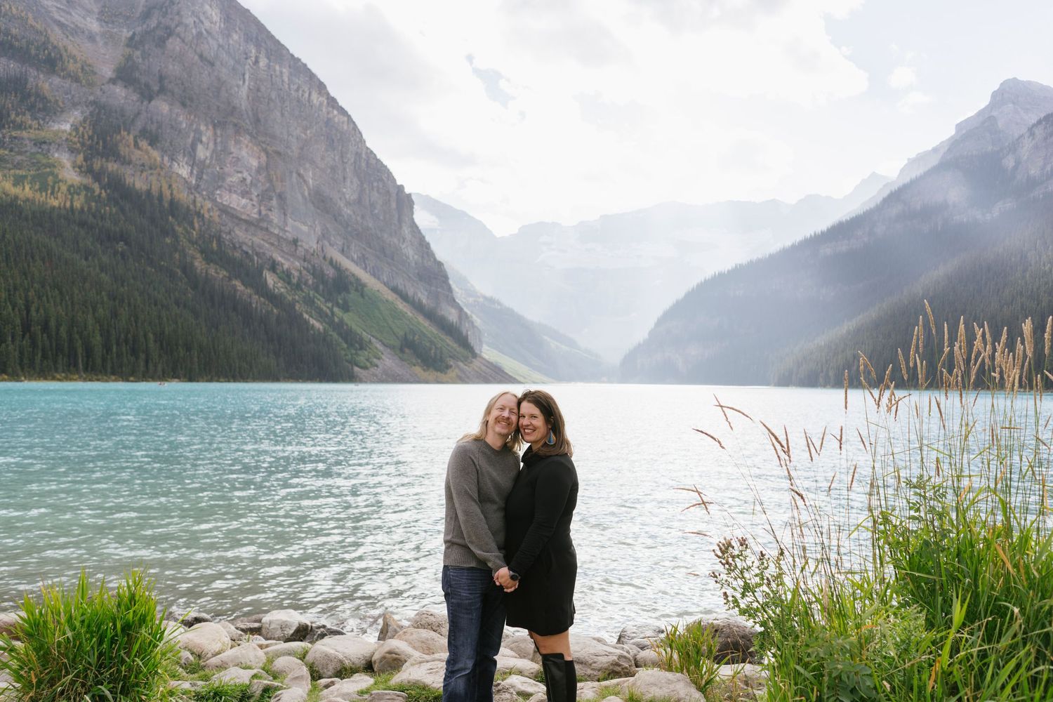 A couple embraces while standing on the rocky shores of Lake Louise in Banff National Park with mountains in the background.