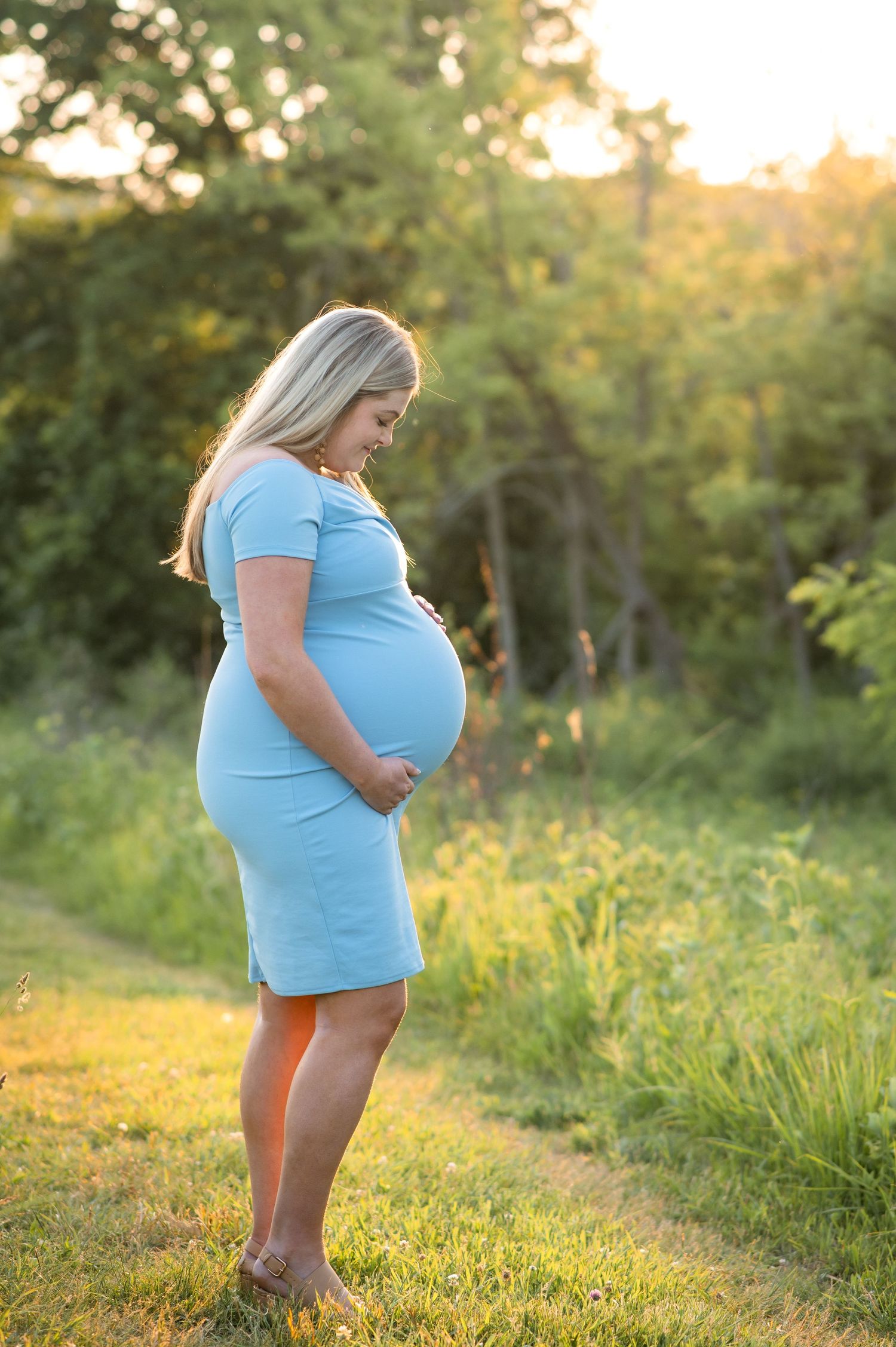 A couple stands in a scenic pine forest path during a maternity photo shoot with sunlight filtering through trees.