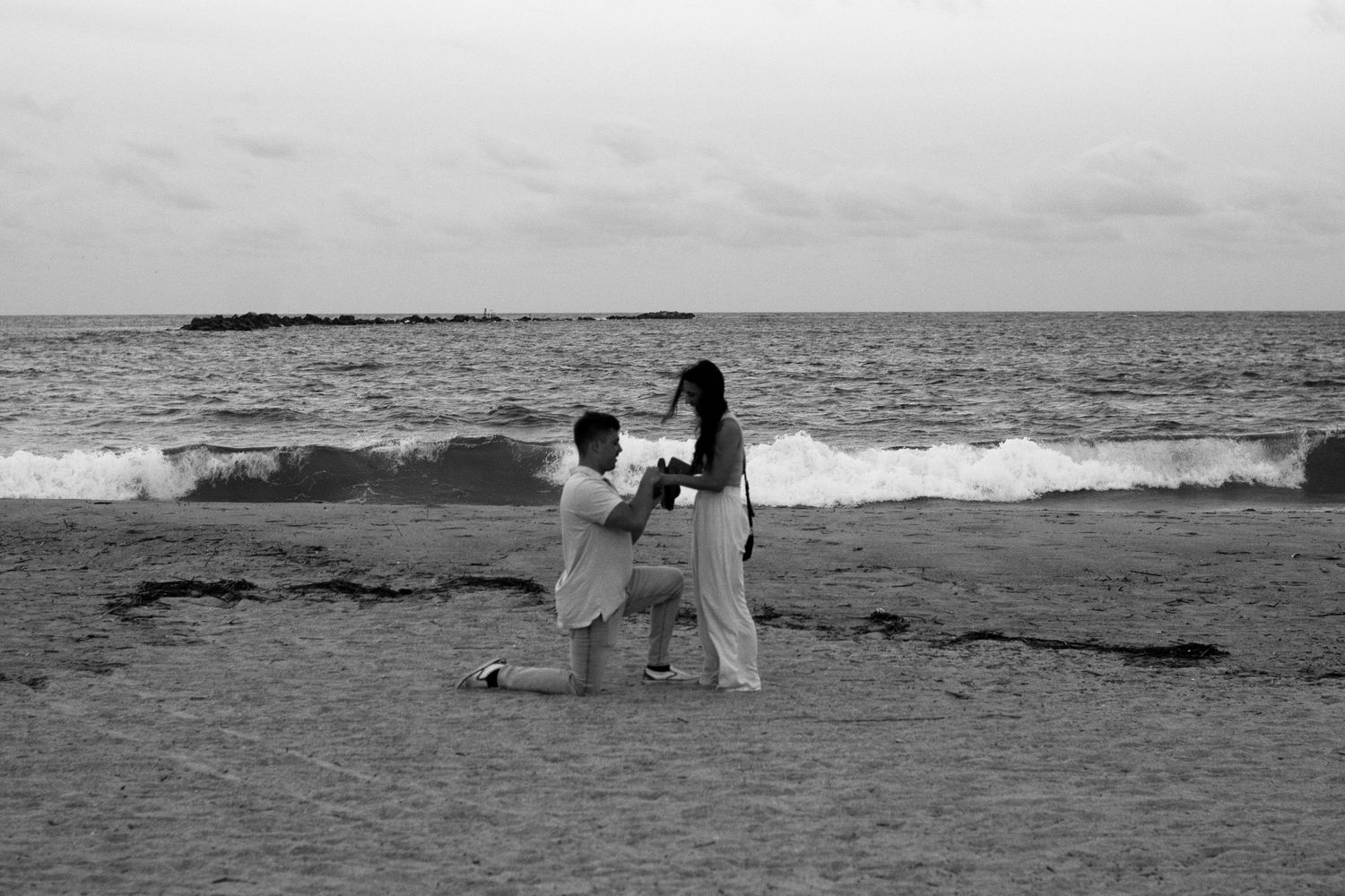 Black and white beach scene of a marriage proposal with waves crashing in the background.