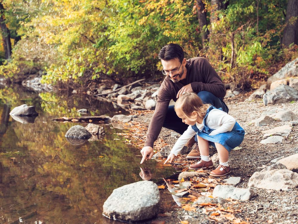 Two people crouch by a peaceful stream, exploring rocks and fallen leaves along the rocky shore at Menotomy Rocks Park in Arlington, MA.