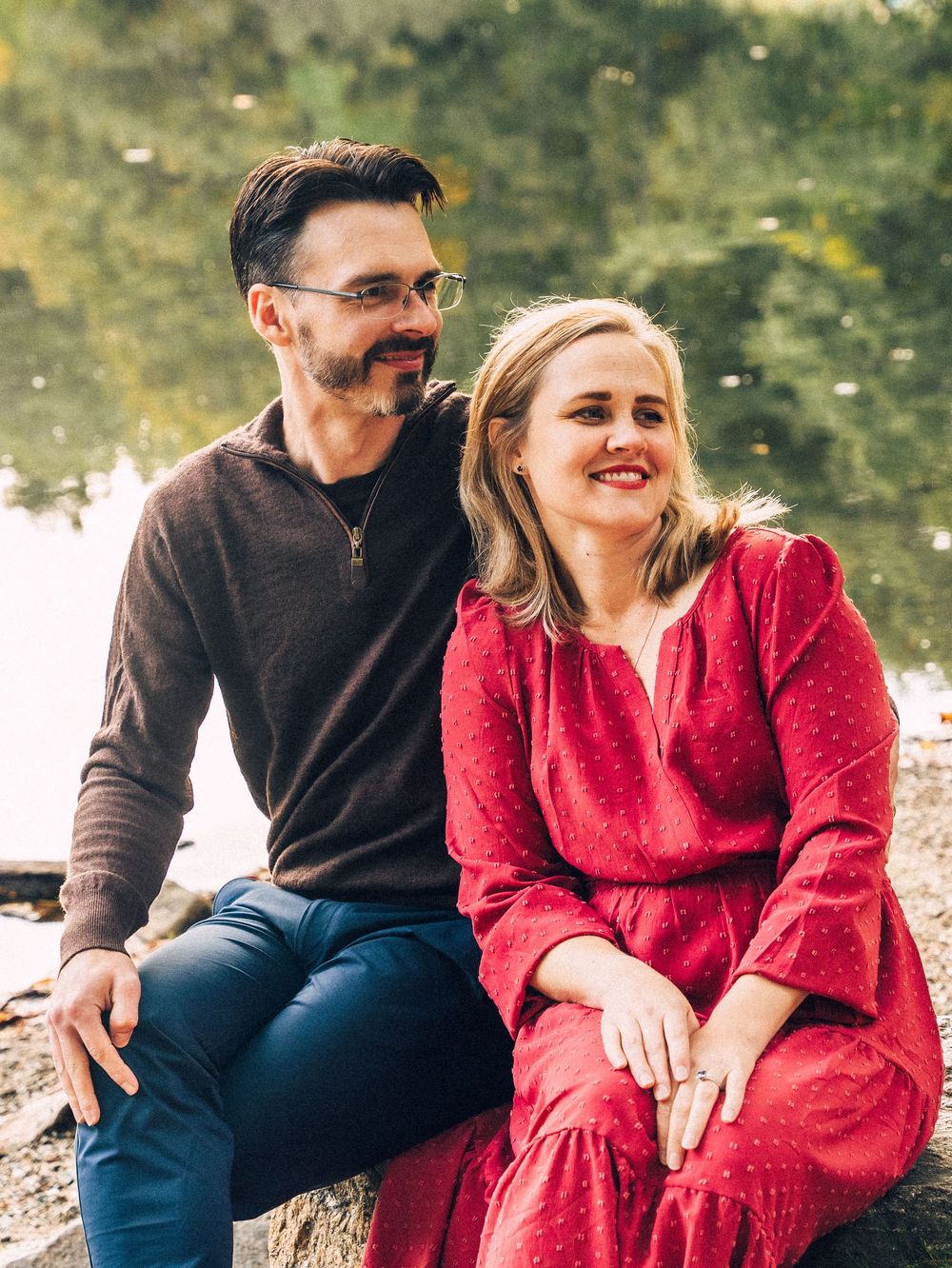A couple in casual attire sits by a waterfront, with one person wearing a red sweater and the other in brown.