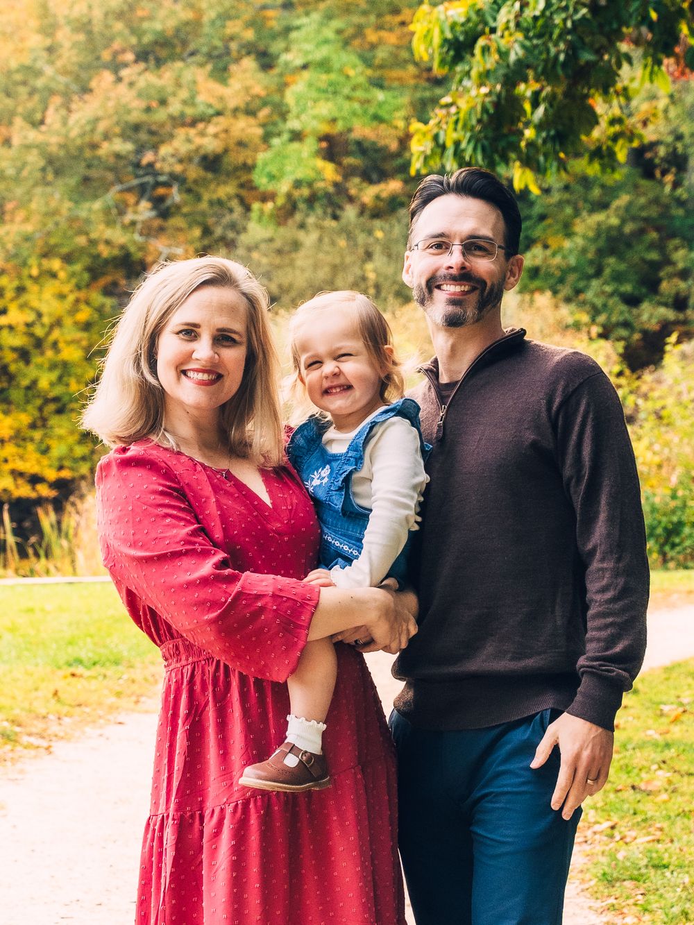 A family wearing red and blue outfits poses together against a backdrop of autumn trees with golden foliage.