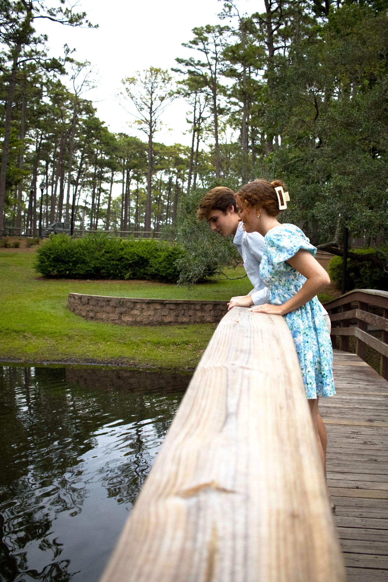 Two people leaning over a wooden railing overlooking a serene pond surrounded by trees.