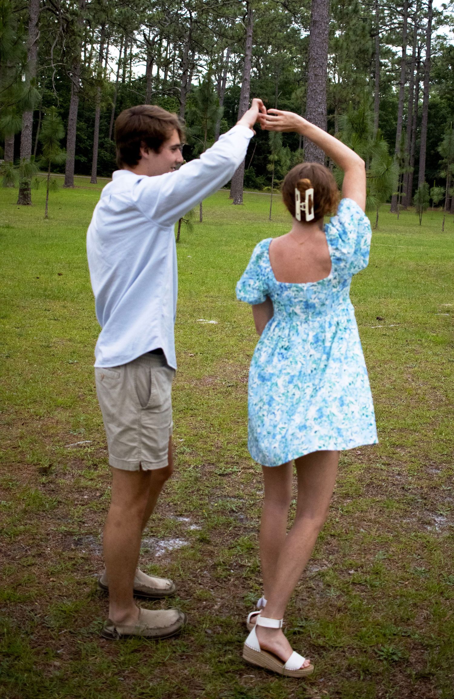 Two people in casual attire dancing playfully on a grassy field with pine trees in the background.