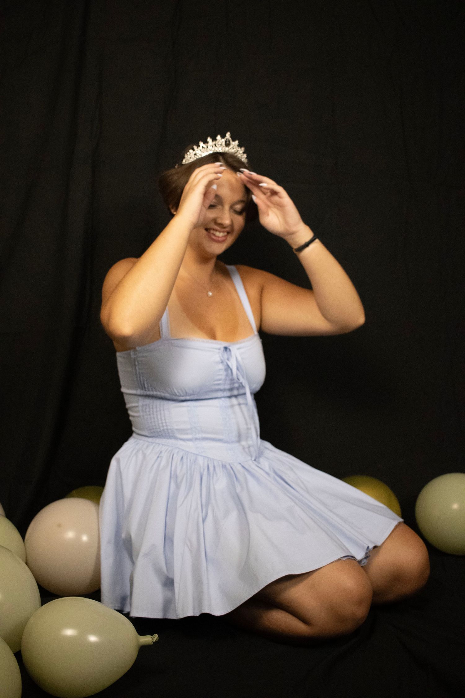 Woman in light blue dress poses playfully against dark backdrop with balloons.
