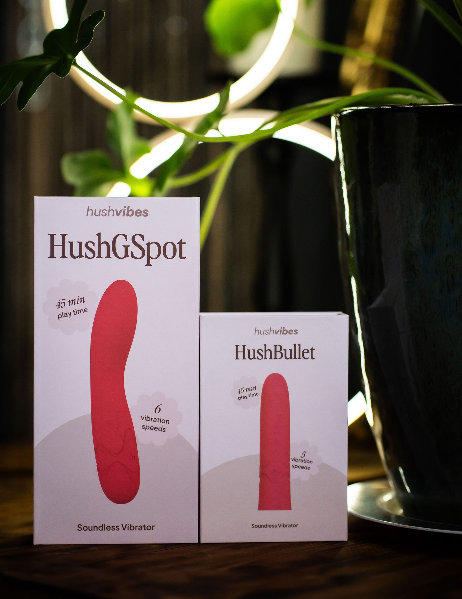 Two pink and white personal massage device product boxes displayed with plants and ring light.